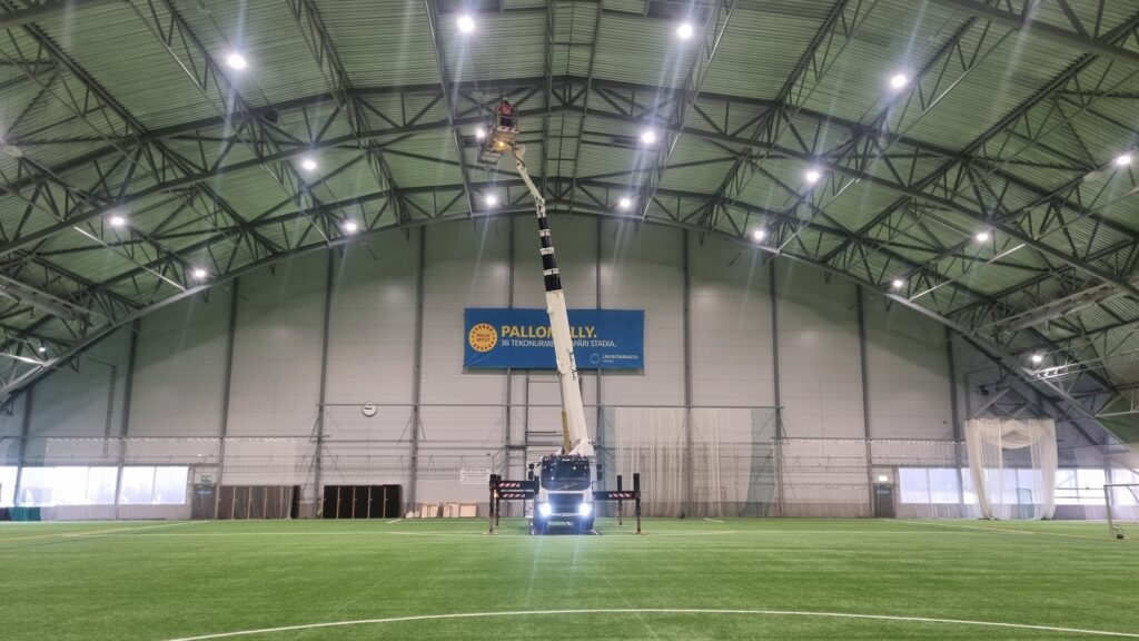 Suuren urheiluhallin sisällä on nostolava-auto, jonka korissa oleva työntekijä tarkastaa hallin kaarevia kattorakenteita.