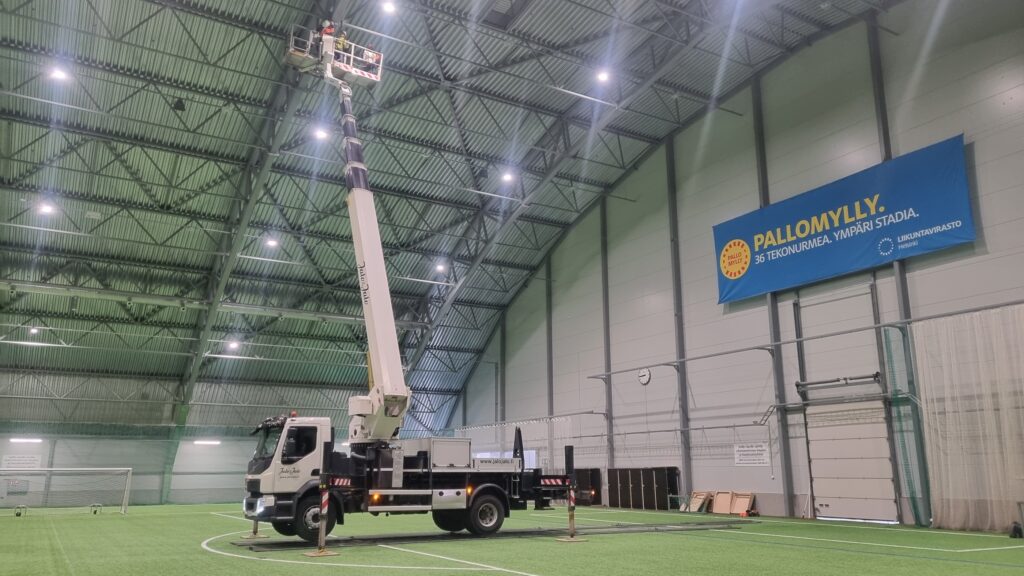 Nostolava-auto palloiluhallin sisätiloissa, korkealla nostokorissa työntekijä tarkistaa kattorakenteita, kuva otettu sivusta