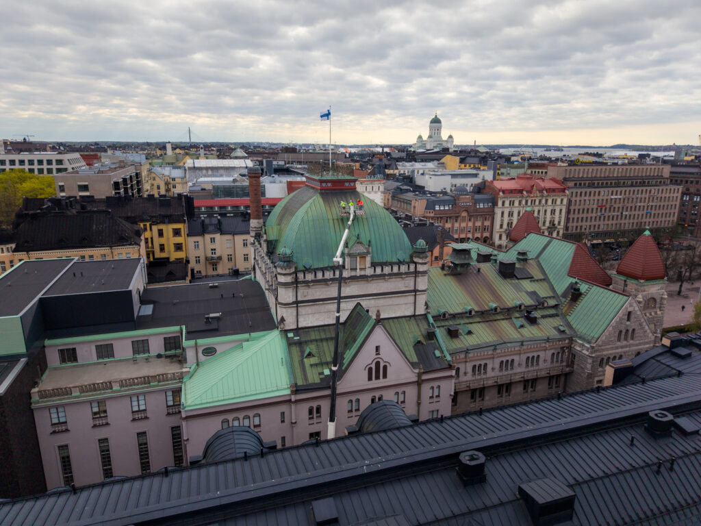 Kattojen yltä kuvattu näkymä, jossa etualalla Kansallisteatterin katto ja yläkerrokset, taustalla Helsinkiä. Kansallisteatterin kattoa tutkitaan nostolava-auton korista.